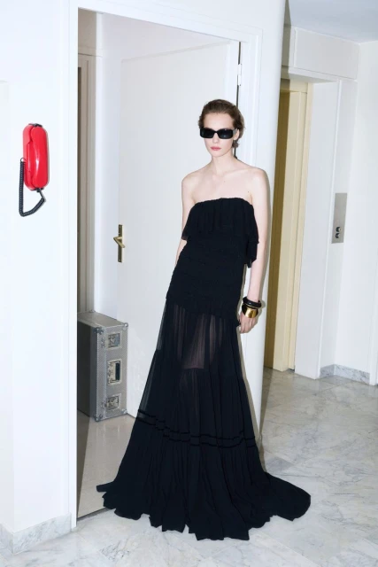 Fashionable adult female posing in a black strapless gown and sunglasses in an indoor setting with a red telephone on the wall.