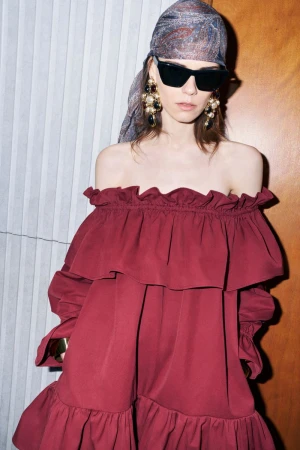 Model wearing an off-the-shoulder burgundy ruffled dress with a headscarf, sunglasses, and large earrings against a textured wall.