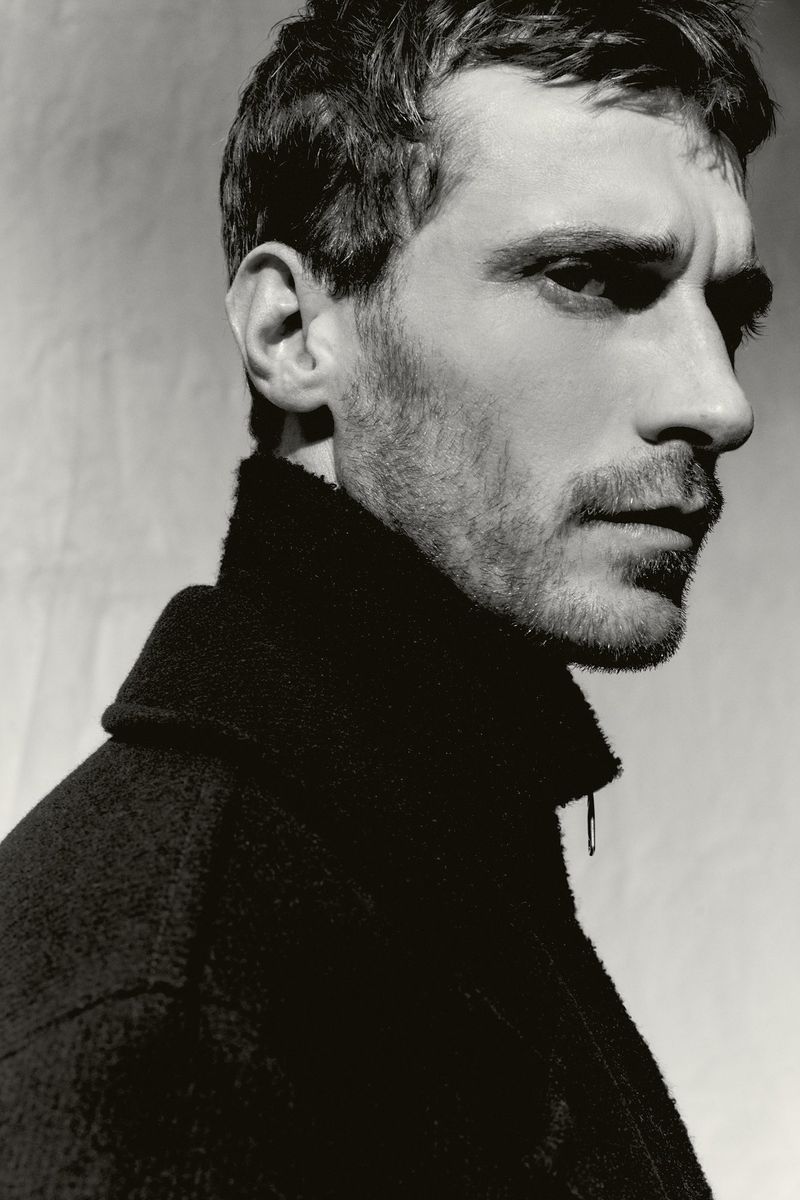Monochrome portrait of a man with short hair and stubble, wearing a black coat, against a blurred, neutral-toned background.