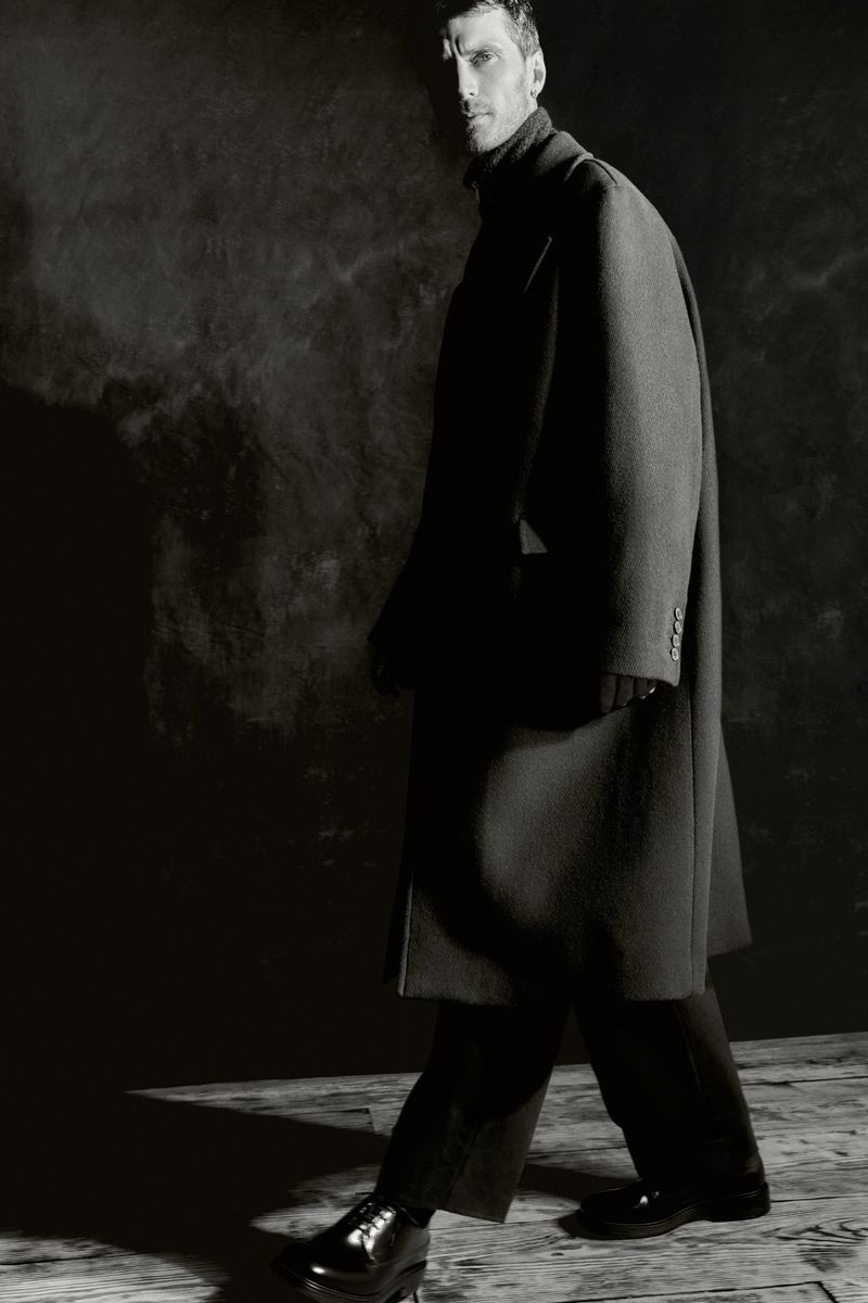 Monochrome portrait of a man in a dark coat and black shoes walking against a mottled gray background, captured in studio lighting.