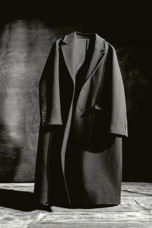 A dark wool overcoat hung against a textured dark background with dramatic studio lighting casting strong shadows.