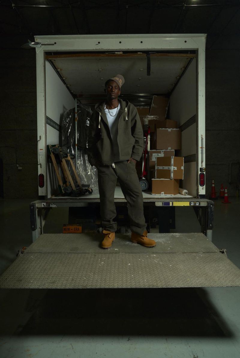An African-American adult male stands on the open back of a white moving truck, with boxes and furniture in the truck's interior.
