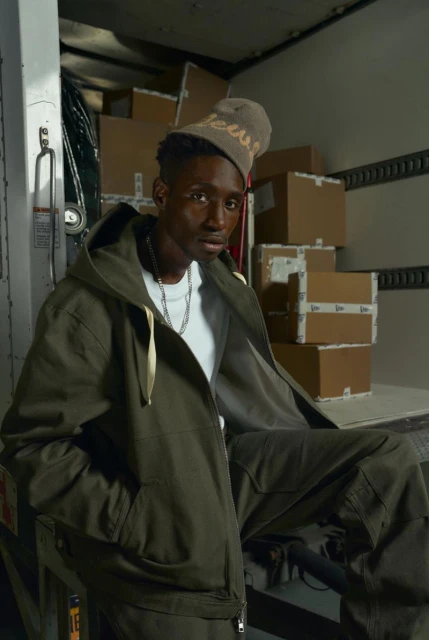 Adult male posing in an olive green jacket and pants sits inside a delivery truck surrounded by stacked cardboard boxes.