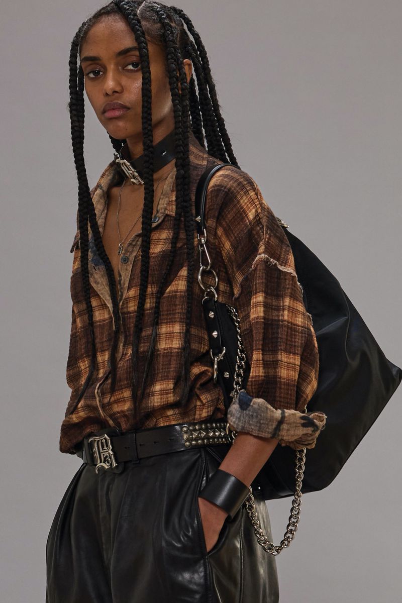 A woman in a brown plaid shirt and black leather pants poses in a studio with a black leather shoulder bag.