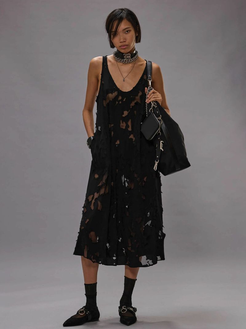 Model in black distressed midi dress holding a dark shoulder bag against a neutral grey studio backdrop.
