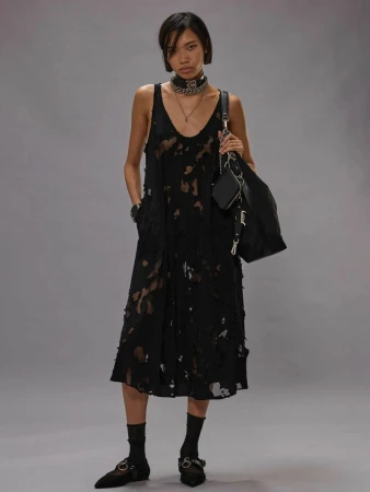 Model in black distressed midi dress holding a dark shoulder bag against a neutral grey studio backdrop.