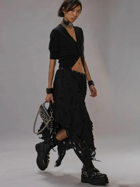 Asian woman walks wearing platform boots, black shredded skirt, cardigan, jewelry, and carrying a chained bag, studio background.