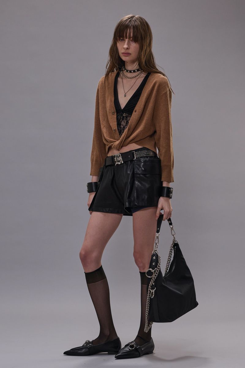 Full-length shot of a woman in a brown cardigan, black shorts, black top, and jewelry standing against a gray background.