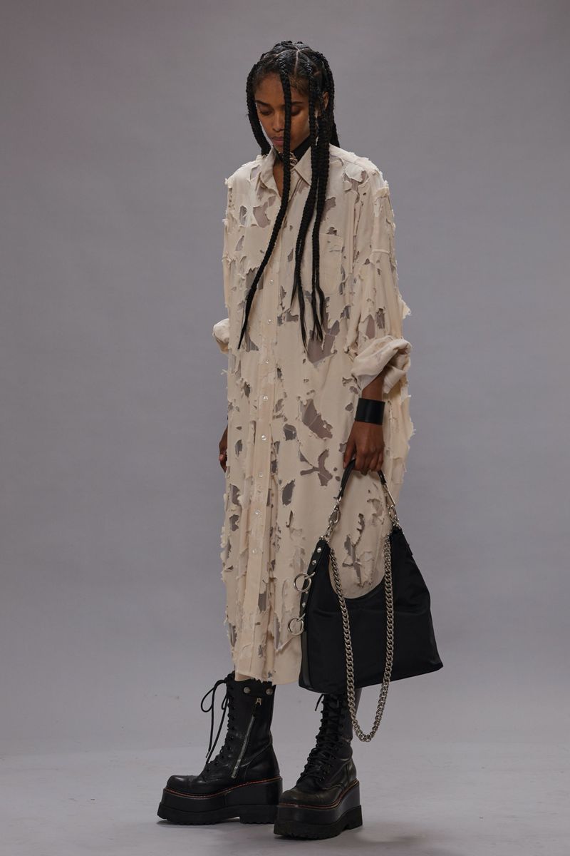 Fashion model posing in a deconstructed shirt-dress, platform boots, and holding a chain purse against a neutral studio backdrop.