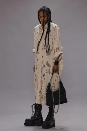 Fashion model posing in a deconstructed shirt-dress, platform boots, and holding a chain purse against a neutral studio backdrop.