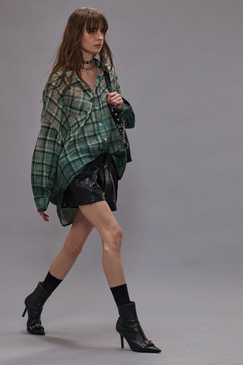 A slender woman strides confidently in a green plaid shirt, black shorts, and heeled boots against a neutral backdrop, carrying a handbag.