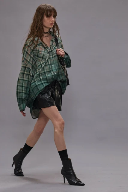 A slender woman strides confidently in a green plaid shirt, black shorts, and heeled boots against a neutral backdrop, carrying a handbag.