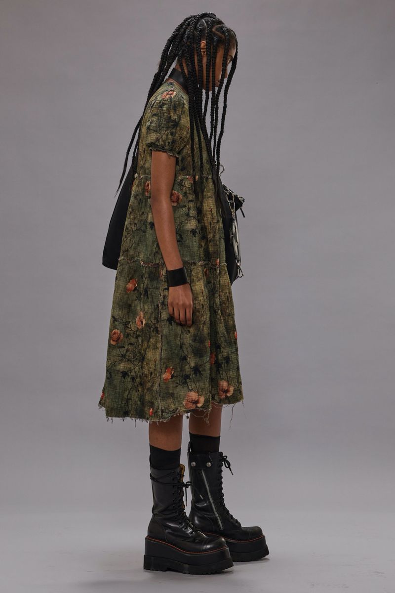 Woman in floral dress, long braids, and platform boots stands in a studio with a neutral background, showcasing a fashion-forward look.