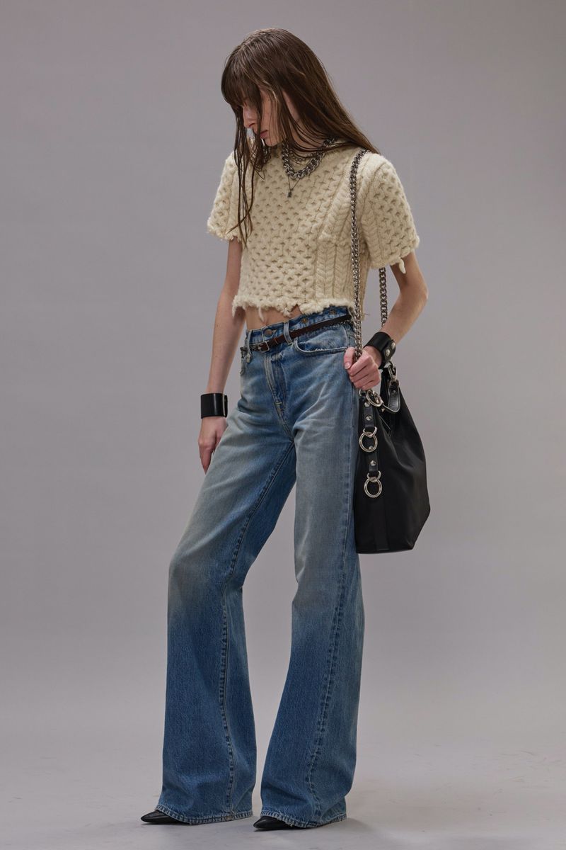 Model wearing a textured cream knit top, distressed blue denim jeans, and carrying a black shoulder bag against a grey studio backdrop.