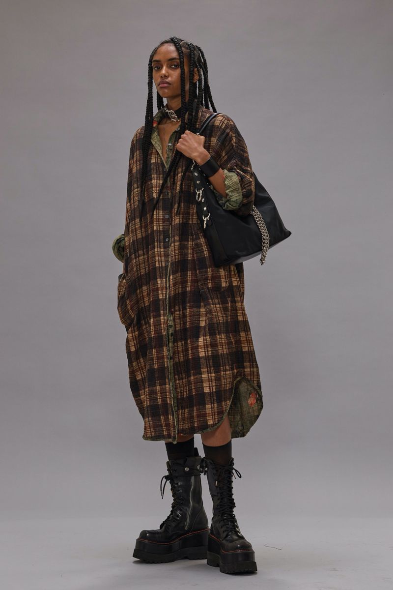 Fashion model with long dark braids poses in a studio wearing a long plaid dress, heavy boots, and shoulder bag against a grey backdrop.