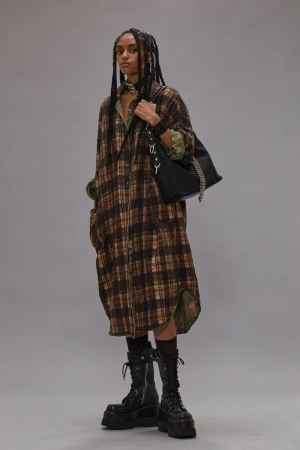 Fashion model with long dark braids poses in a studio wearing a long plaid dress, heavy boots, and shoulder bag against a grey backdrop.