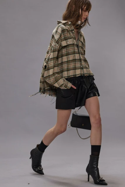 A woman walks in a studio, wearing a plaid shirt and leather shorts with black heeled boots and a black purse on a chain.