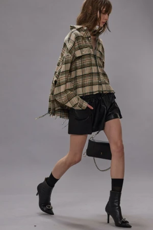 A woman walks in a studio, wearing a plaid shirt and leather shorts with black heeled boots and a black purse on a chain.