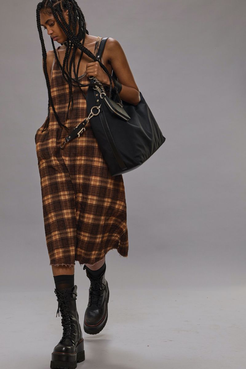 An adult woman with long braids models in a studio, wearing a plaid dress, black socks, and black platform boots, holding a black bag.