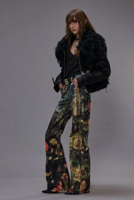 A woman poses in printed pants, a black top, and a furry-collared leather jacket against a gray background in a fashion studio shot.