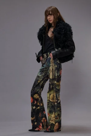 A woman poses in printed pants, a black top, and a furry-collared leather jacket against a gray background in a fashion studio shot.