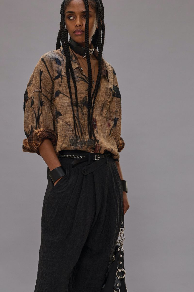 A woman poses in a brown floral print top and black pants, accessorized with a belt and bracelets, against a neutral backdrop.