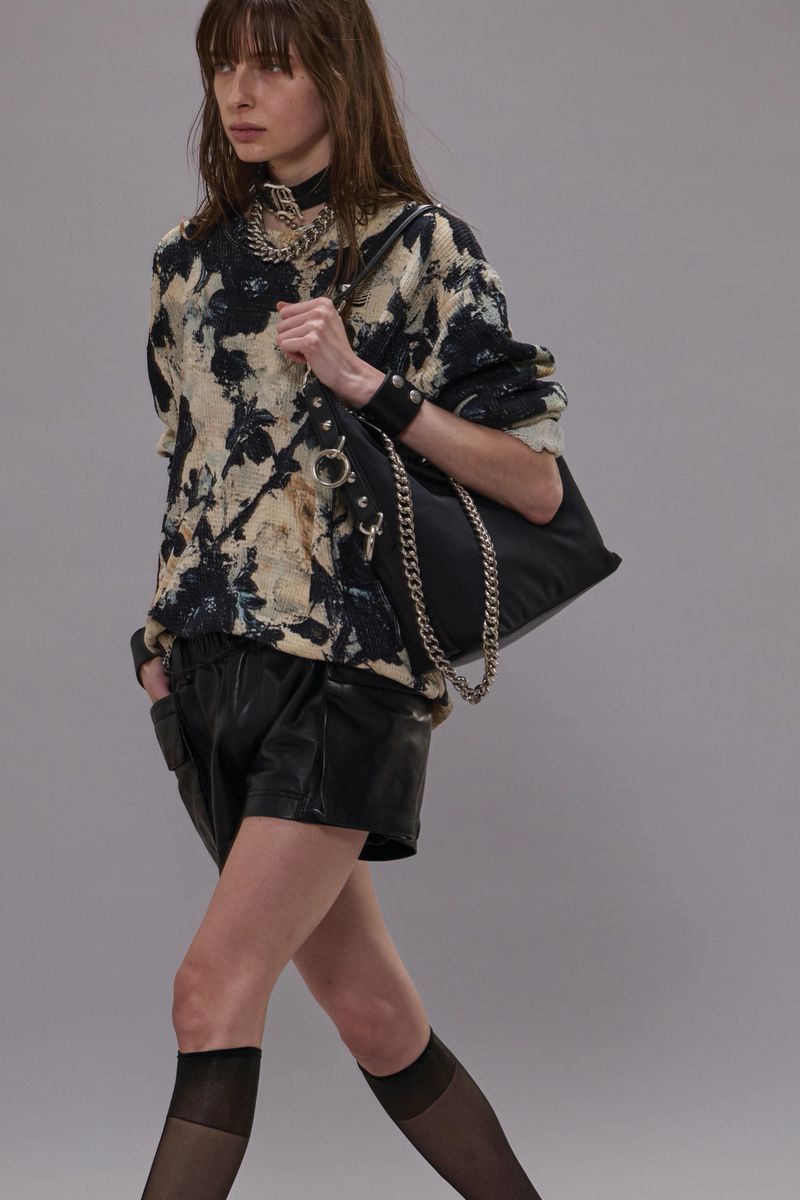 An adult female model walks in a studio wearing a printed sweatshirt, black shorts, tights, jewelry, and a shoulder bag with a chain.