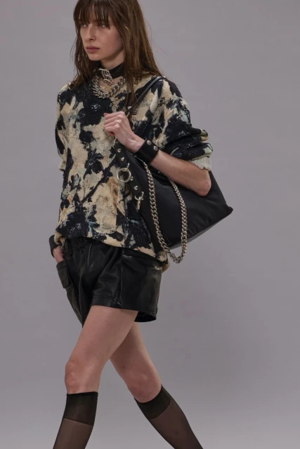 An adult female model walks in a studio wearing a printed sweatshirt, black shorts, tights, jewelry, and a shoulder bag with a chain.