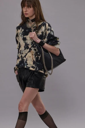 An adult female model walks in a studio wearing a printed sweatshirt, black shorts, tights, jewelry, and a shoulder bag with a chain.