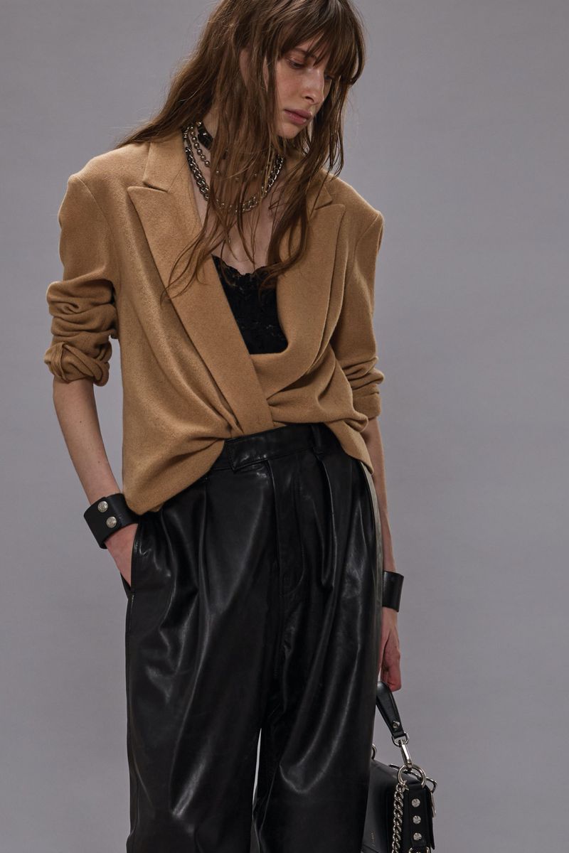 Fashionable woman in a tan blazer and black leather pants, standing in front of a gray background, showcasing modern street style.