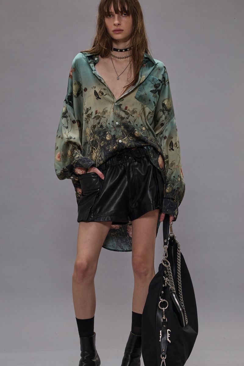 A woman poses in an unbuttoned floral shirt, black shorts, black boots and a matching purse against a neutral backdrop.