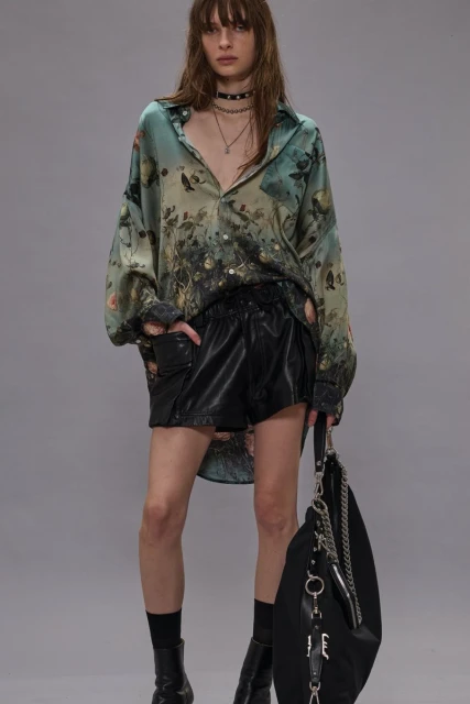 A woman poses in an unbuttoned floral shirt, black shorts, black boots and a matching purse against a neutral backdrop.