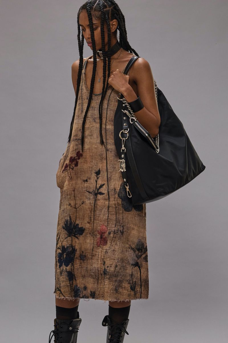 A tanned-skin woman wearing a brown floral dress and boots with braided hair holds a black purse against a gray studio backdrop.