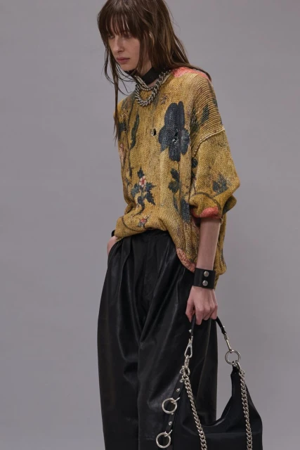 Fashion portrait of a woman with bangs wearing a floral sweater, leather pants, chain jewelry, and holding a black chain purse.