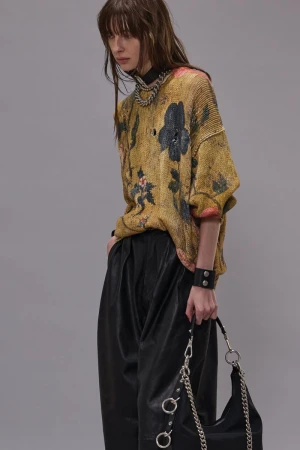Fashion portrait of a woman with bangs wearing a floral sweater, leather pants, chain jewelry, and holding a black chain purse.