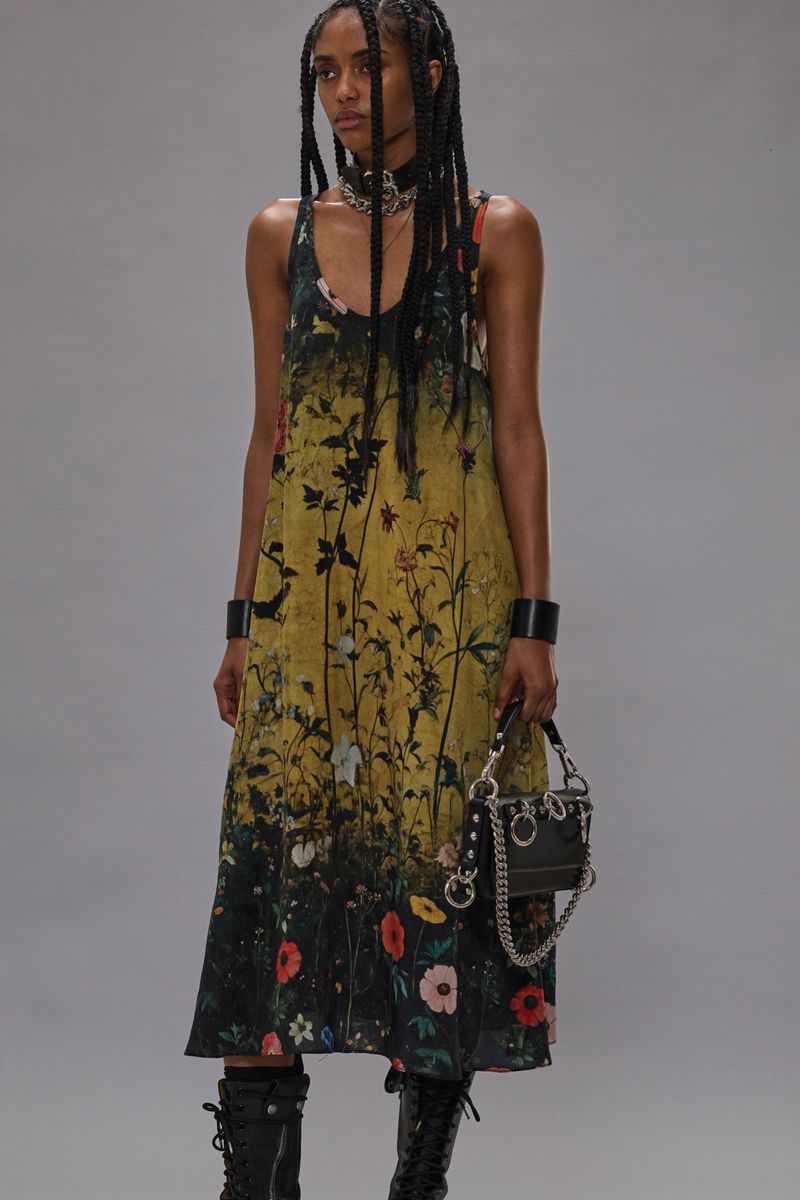 Woman in floral-print dress, choker, and boots holding a small, studded purse on a grey backdrop. Braided hair, wrist cuffs.