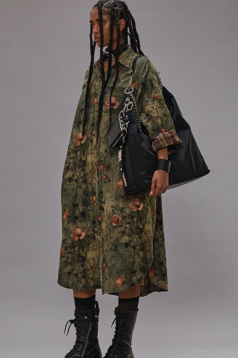 A stylish adult woman wearing a floral dress and combat boots stands in a studio with a black shoulder bag and braids.