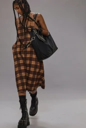 Model in a brown plaid dress and black platform boots carrying a large black shoulder bag against a grey background