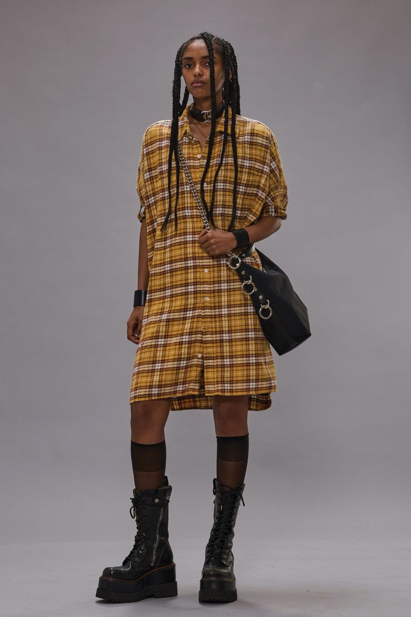 Model in yellow plaid shirt dress wearing platform boots and carrying a black shoulder bag against a grey background