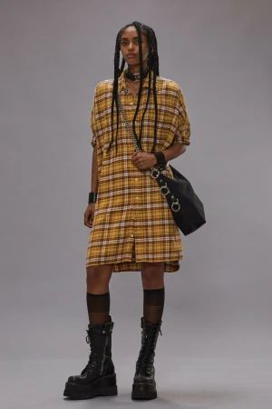 Model in yellow plaid shirt dress wearing platform boots and carrying a black shoulder bag against a grey background