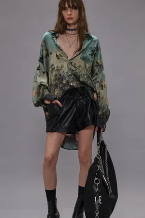 Model in printed silk shirt and black leather shorts holding a large bag against a grey seamless backdrop for R13 Pre-Fall 2025.