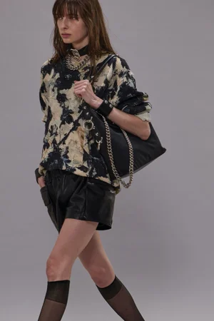Model walking in a printed top and black shorts carrying a shoulder bag against a grey seamless backdrop for R13 Pre-Fall 2025 Lookbook