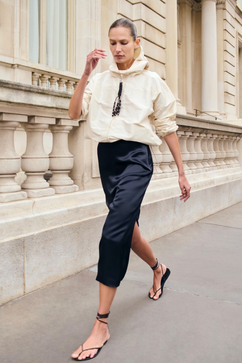 Fashionable woman walking in an urban environment, wearing a cream-colored jacket, black skirt, and flat sandals in a street setting.
