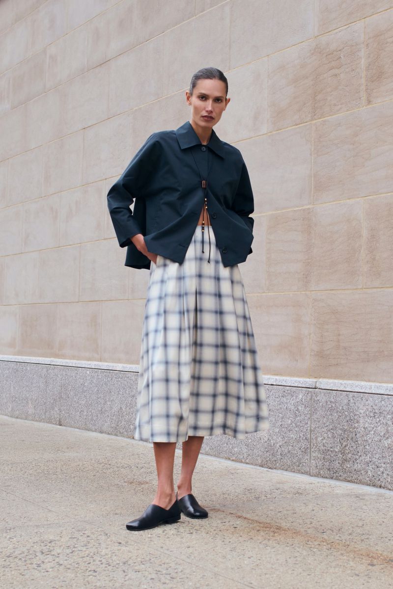 Woman poses in a navy jacket, plaid midi skirt, and black flats against an exterior wall, showcasing fashion and style.