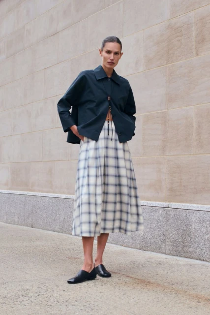 Woman poses in a navy jacket, plaid midi skirt, and black flats against an exterior wall, showcasing fashion and style.