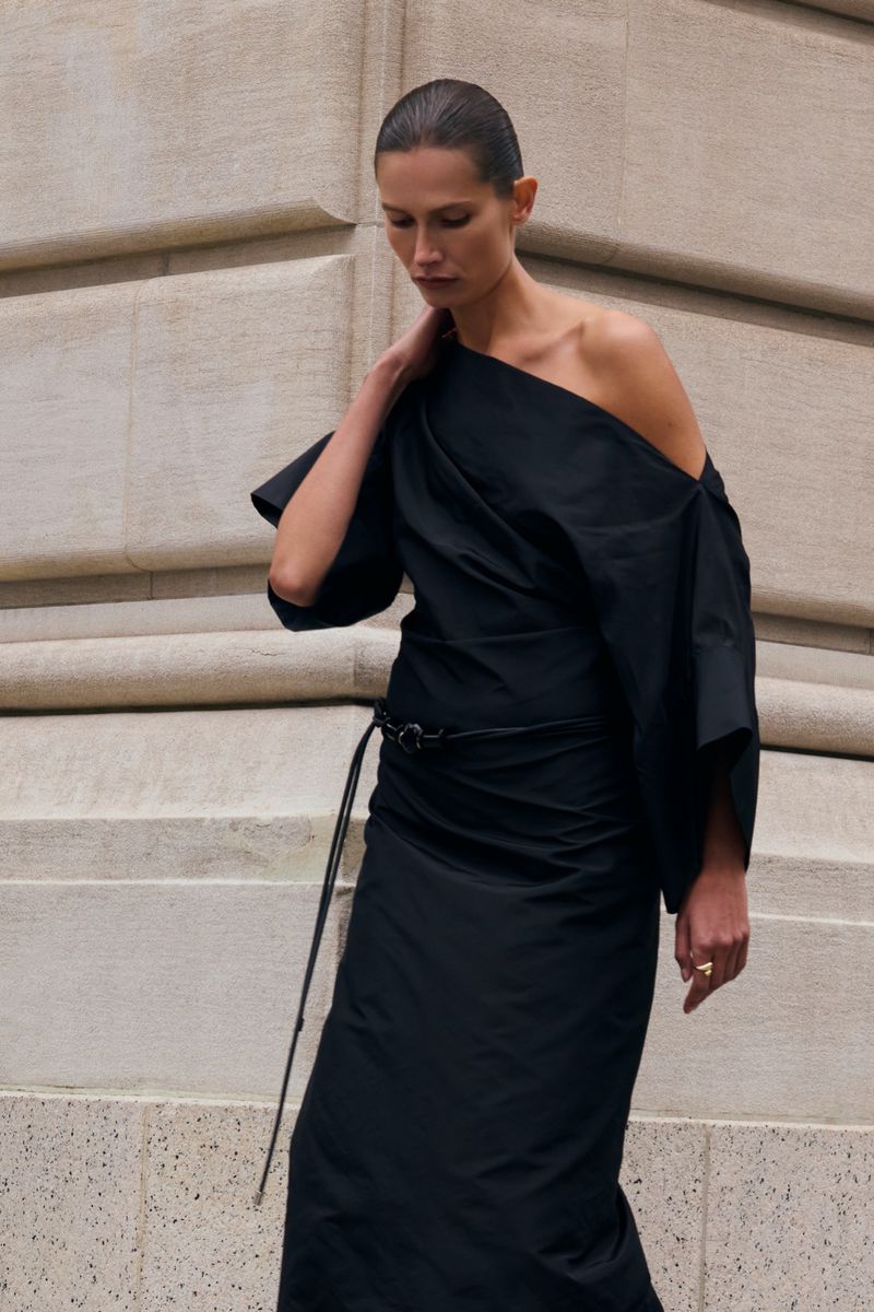 Model in a black one-shoulder dress standing against a structured stone building exterior for Kallmeyer pre-fall 2025 lookbook.