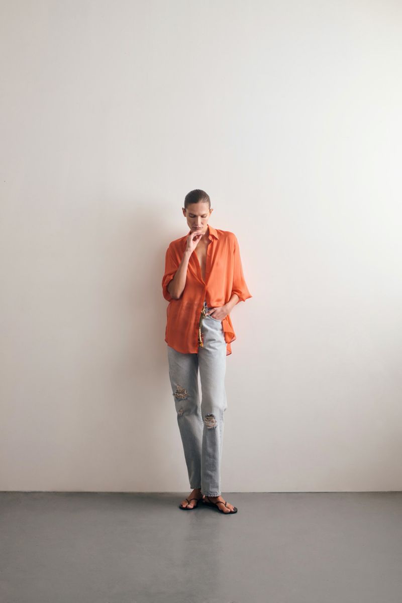 Full-length shot of a woman in a coral button-down shirt and light wash ripped jeans posing against a plain white wall.