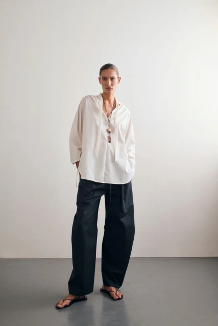 Fashion shot of a woman in a white oversized shirt and black pants standing in front of a white wall in a minimalist style.
