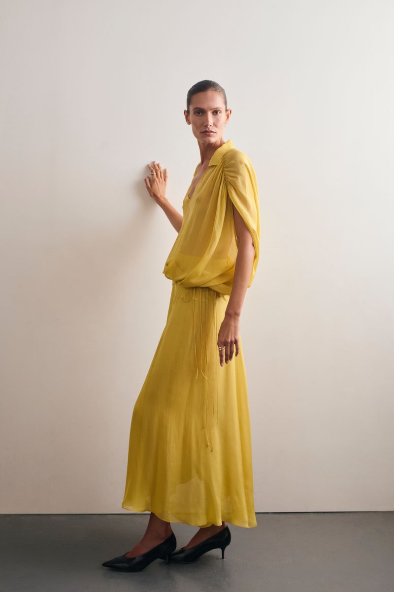 Elegant adult woman in yellow chiffon dress and black heels strikes a pose against a neutral wall in a fashion photography studio setting.