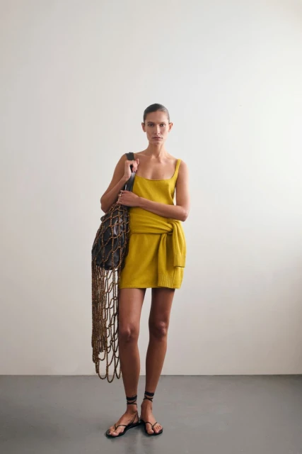 A woman poses indoors wearing a yellow dress and black sandals, holding a dark bag with a light brown net design, against a neutral backdrop.